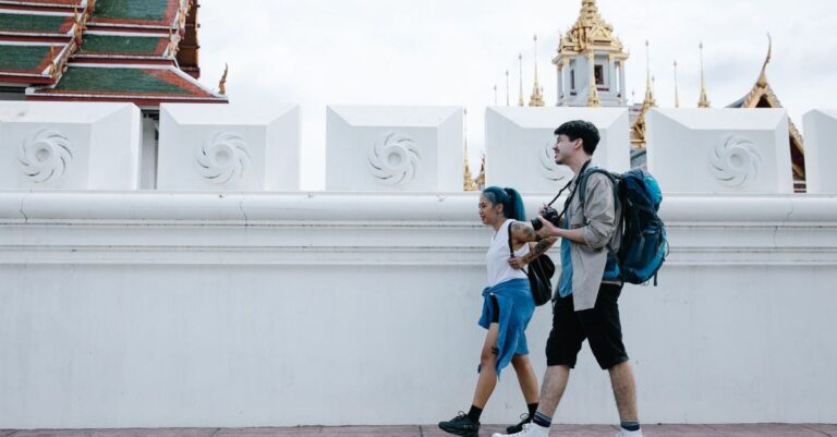 タイの寺院の塀沿いを、小さなバッグとリュックだけで歩く2人の旅行者